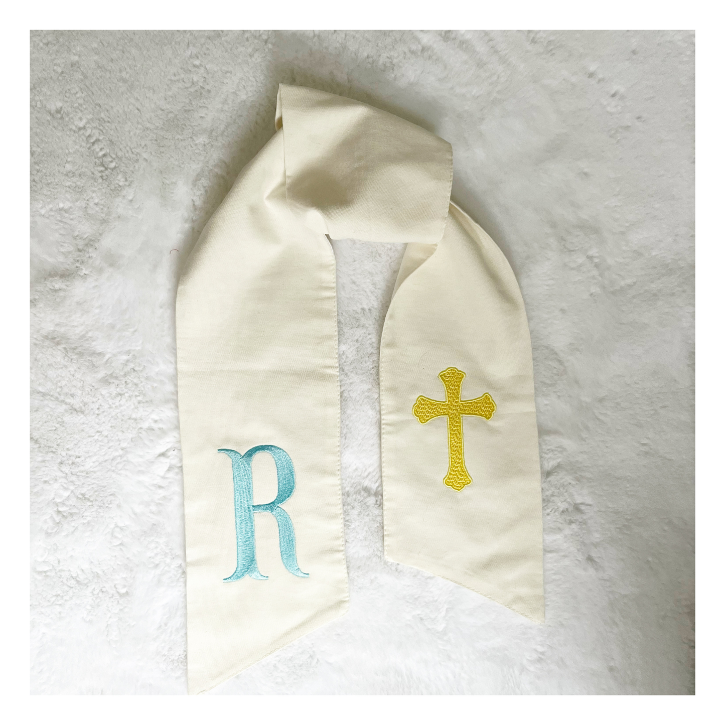 Embroidered white wreath sash with blue R in fishtail monogram font on one side and yellow cross on other side. 