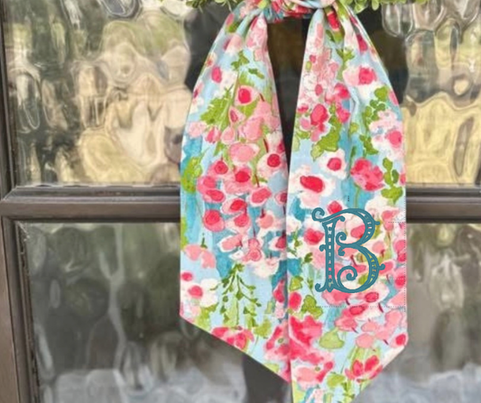 Floral wreath sash with a monogram 'B' in fancy font hanging on a glass door.