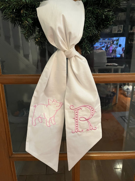 A white sash with a embroidered outlined pig design and the letter 'R' monogrammed on it in a whimsical font with dots and curves.