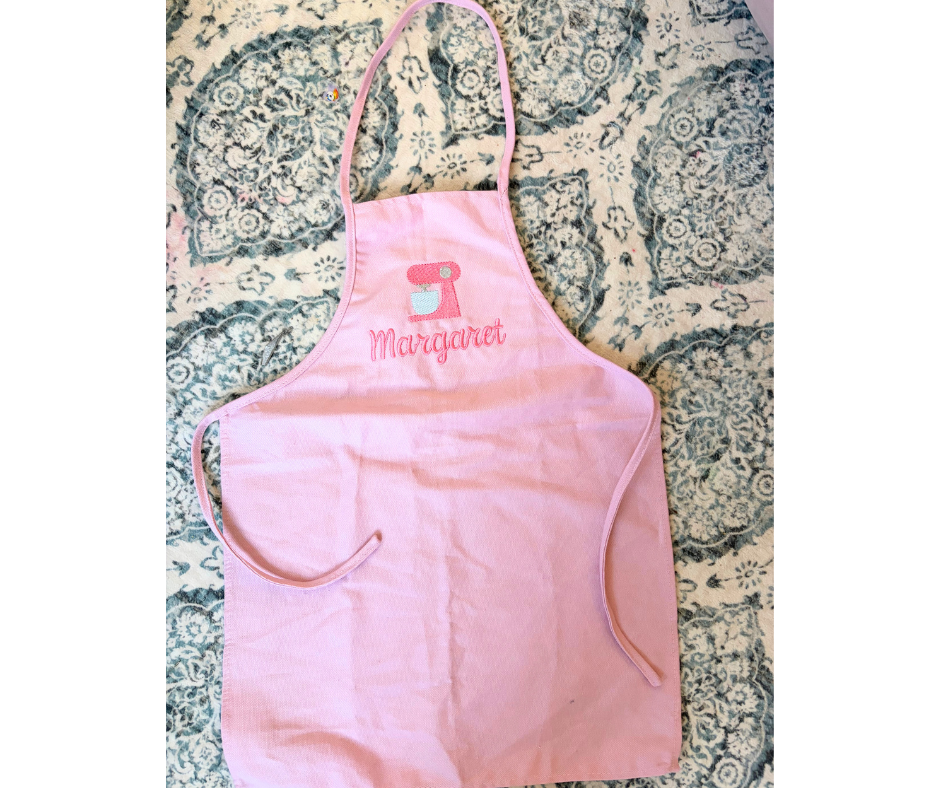 Pink apron with embroidered name and mixer design on a patterned fabric background
